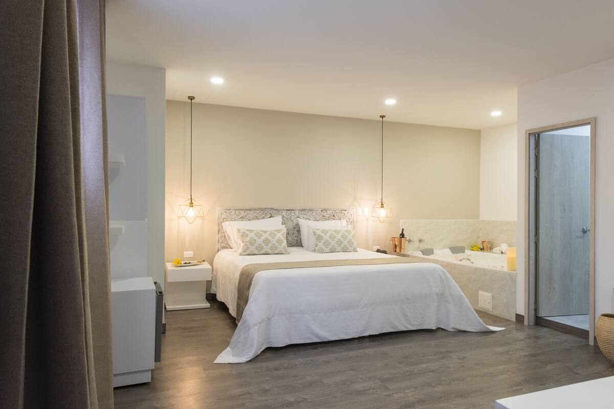 A modern bedroom with a large bed, bedside tables, a built-in bench, a vanity with a mirror, and a shower area.