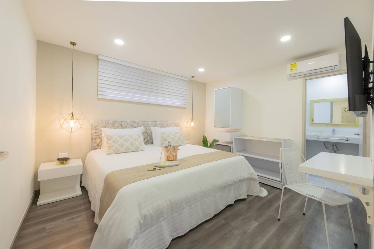 A modern hotel room with a neatly made bed, a small desk with a chair, a TV, and a bathroom with a mirror and sink.