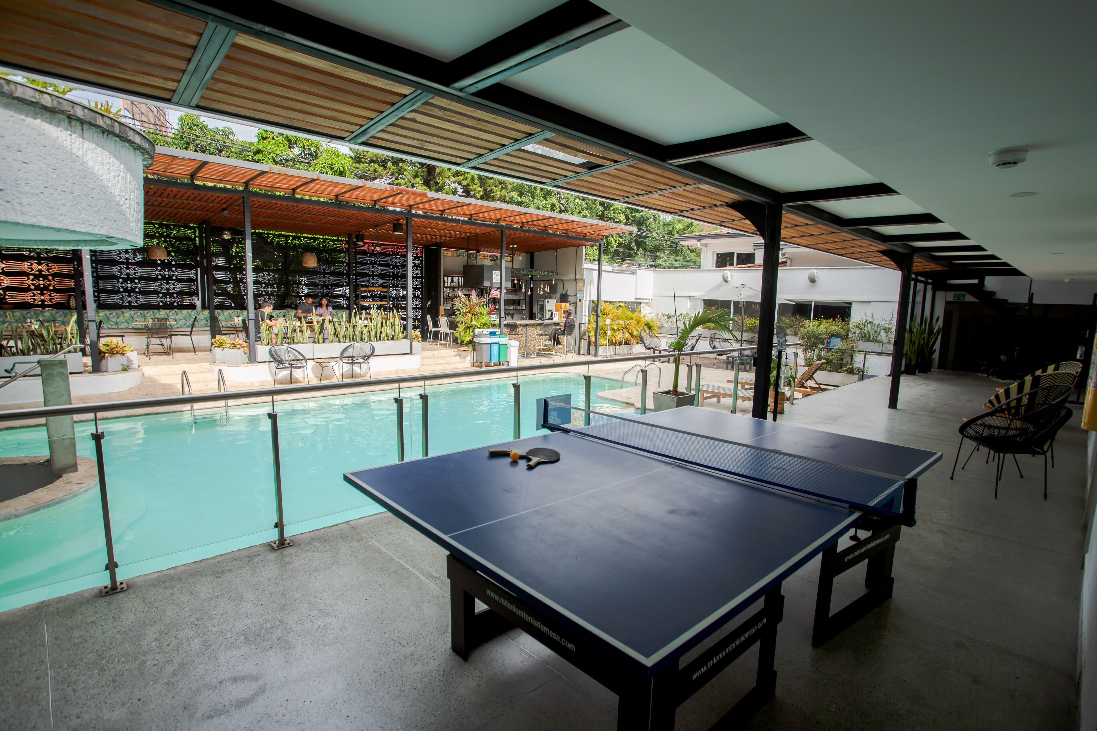 An outdoor pool area with a ping pong table, chairs, and a bar area with plants and umbrellas.
