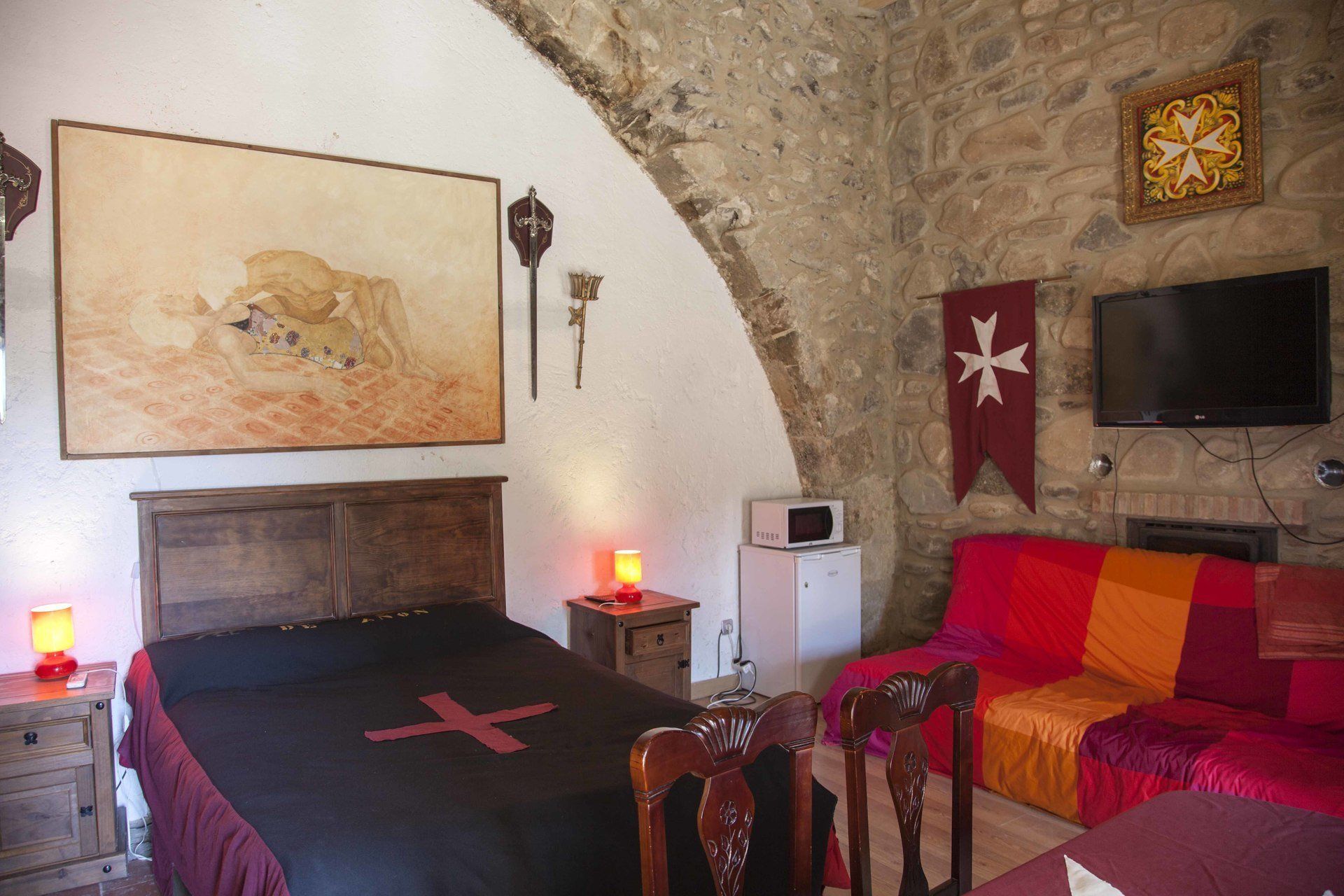 Une pièce avec un mur en pierre, un lit recouvert d’une couverture à croix rouge, une tête de lit en bois, une petite table avec une lampe, un téléviseur et un drapeau accroché au mur.