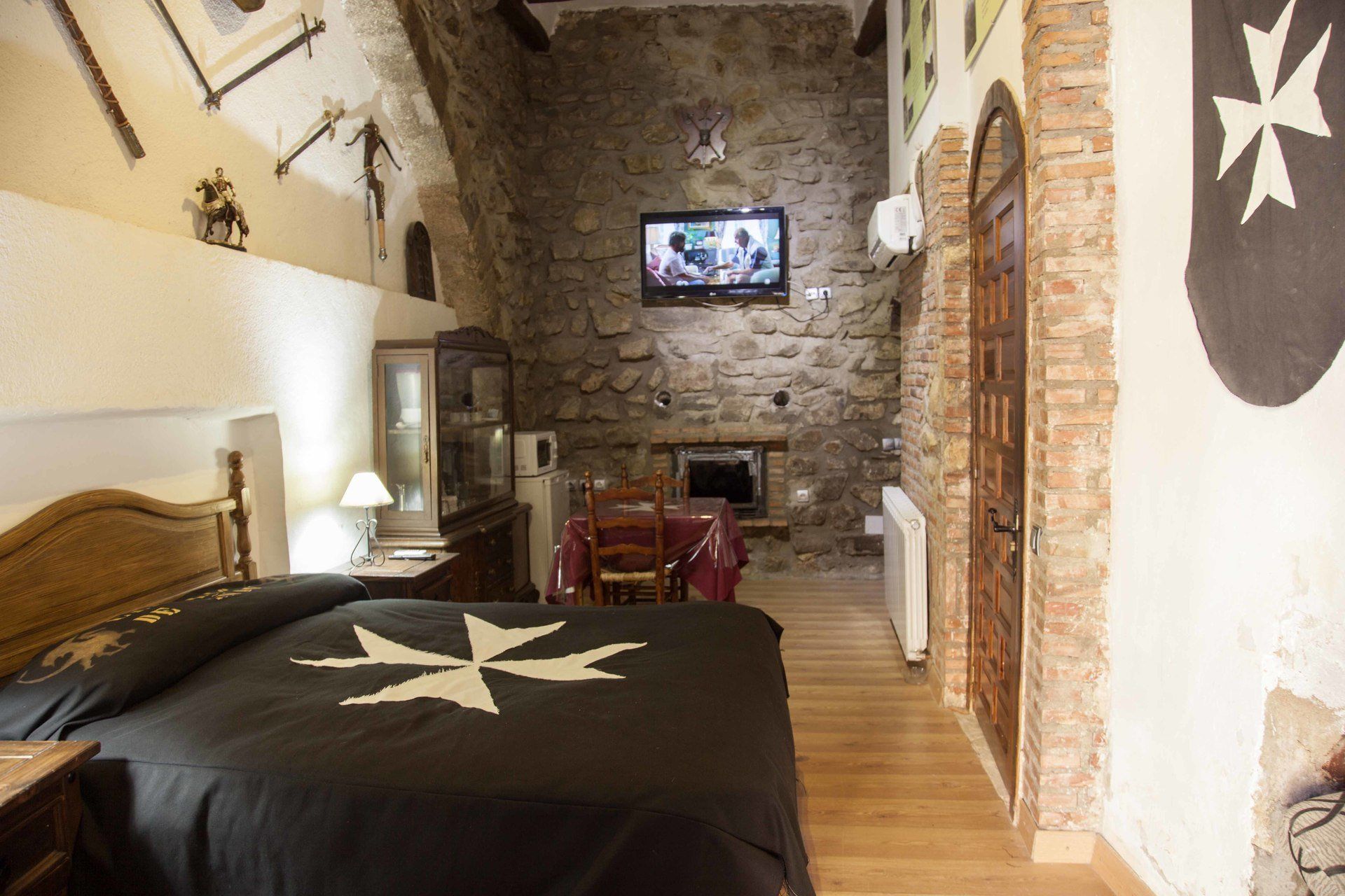 Un dormitori amb paret de pedra, televisió penjada a la paret, llit amb edredó negre, capçalera de fusta, tauleta de nit de fusta, làmpada, armari i una llar de foc.