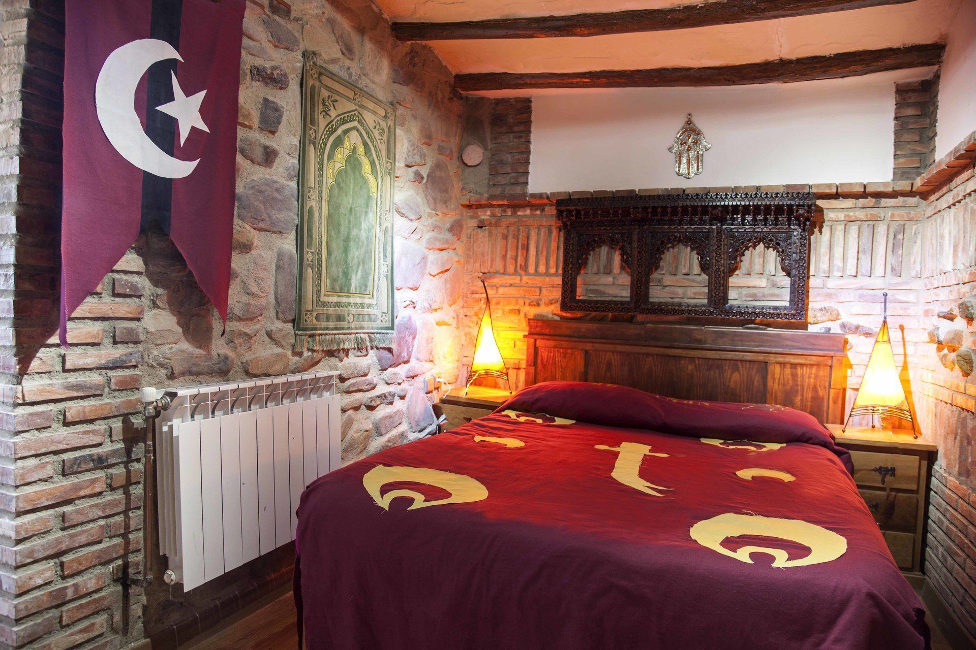 Une chambre avec un mur en pierre, un lit recouvert d’une couverture rouge, deux lampes fixées au mur et un drapeau suspendu.