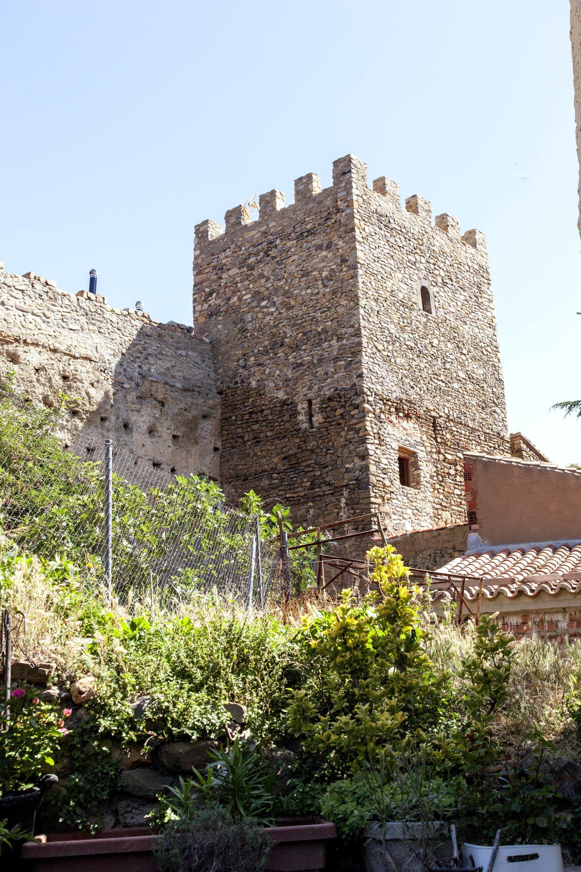 A stone tower with a crenellated top, surrounded by vegetation and a metal fence.