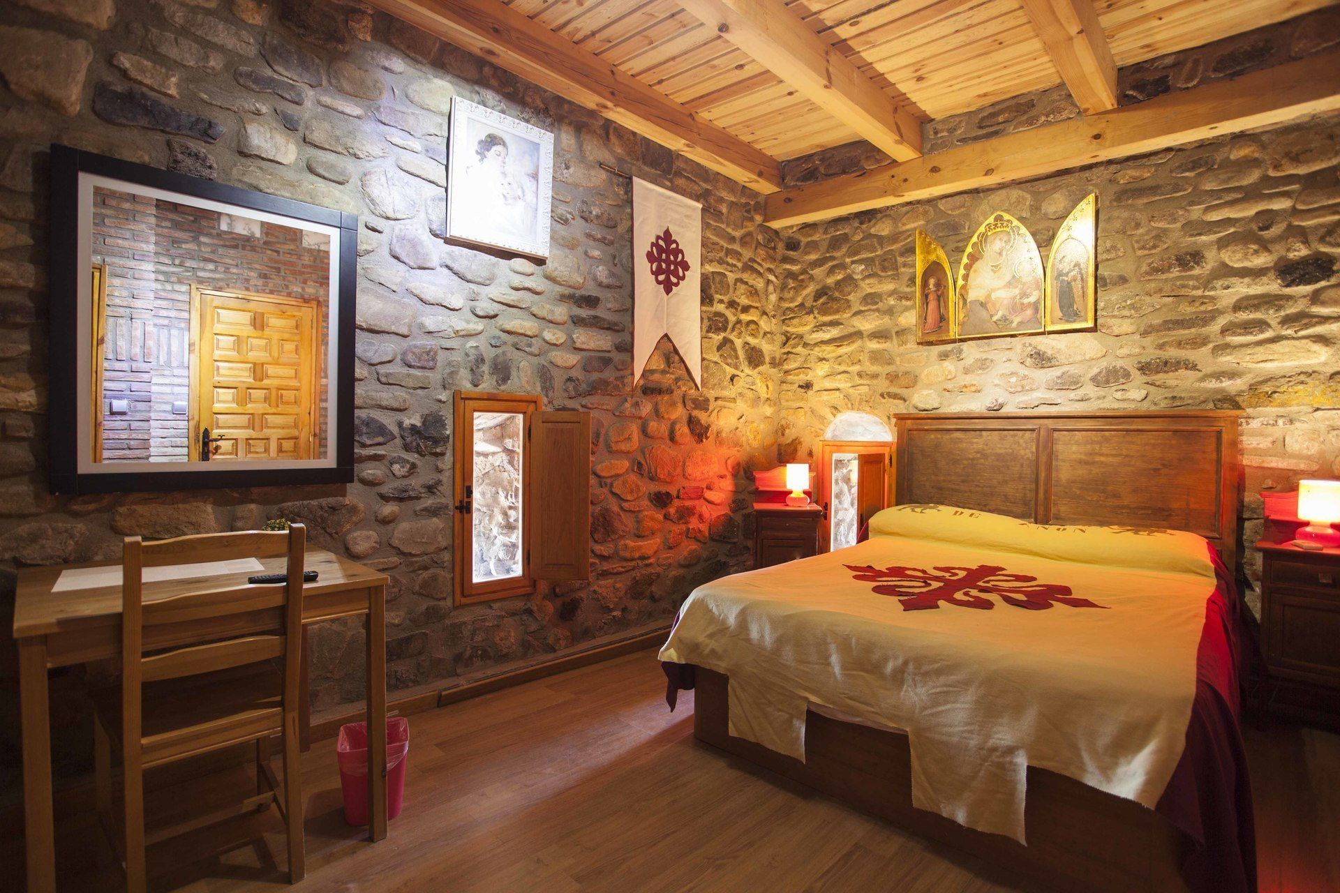 A rustic room with a stone wall, wooden ceiling beams, a bed with a white bedspread, a wooden chair, and a small table with a lamp.
