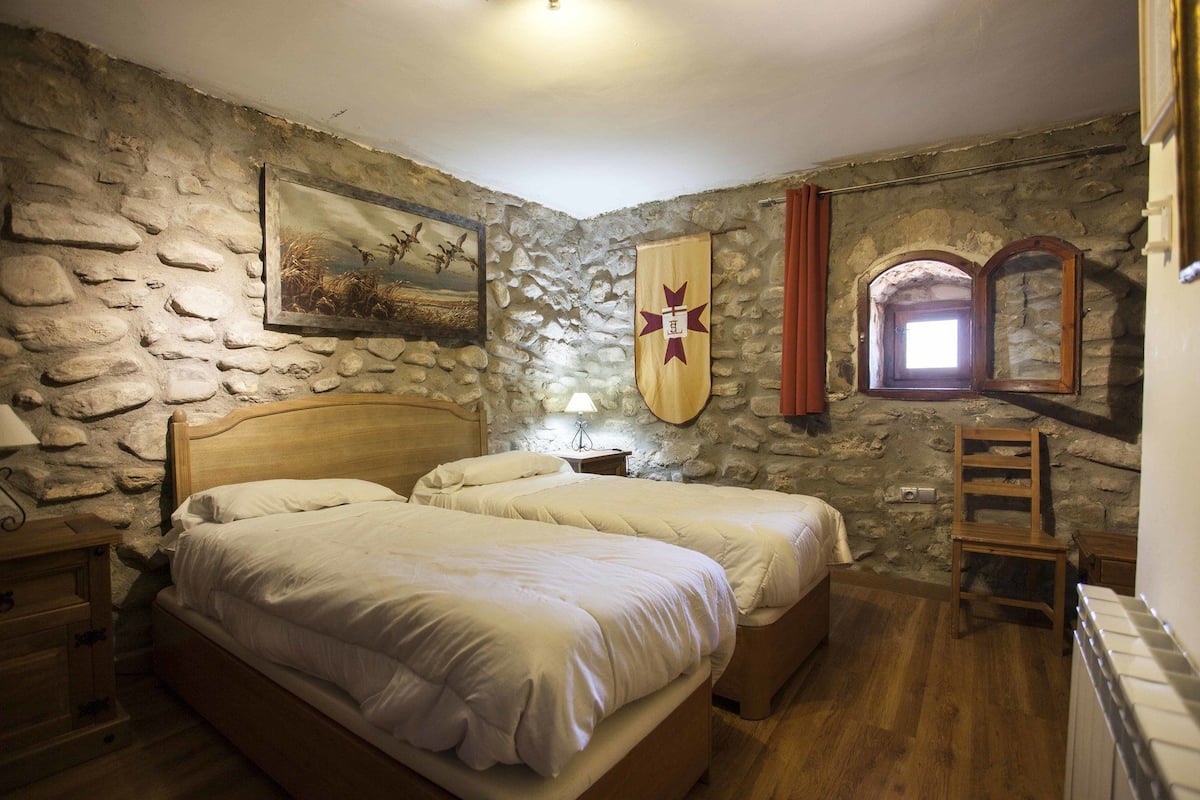 Un dormitorio con pared de piedra, una cama, una mesita de noche, una lámpara, una escalera de madera y una ventana con cortinas.