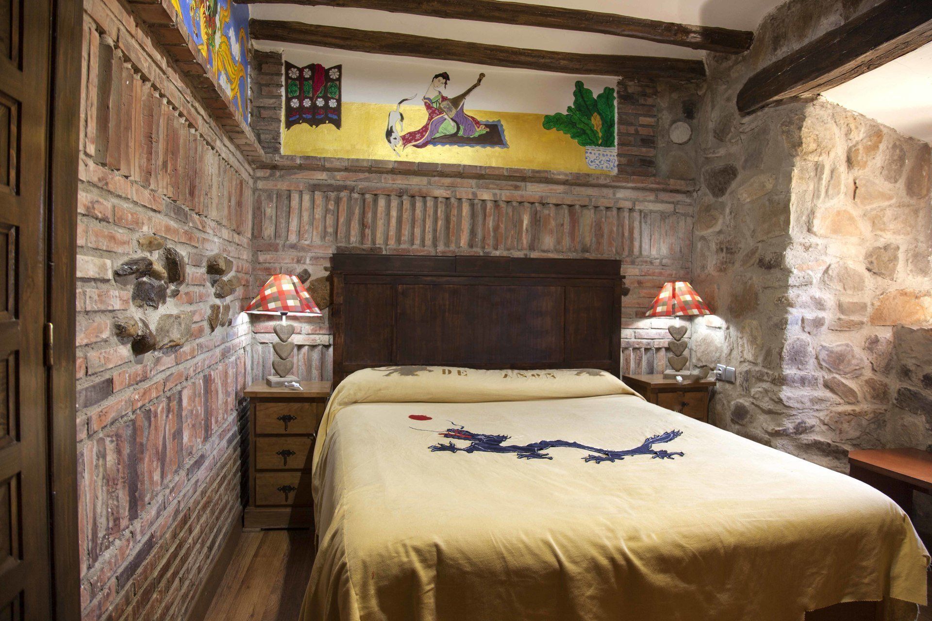A rustic bedroom with a wooden headboard, a bed with a decorative bedspread, stone walls, and a mural on the wall.
