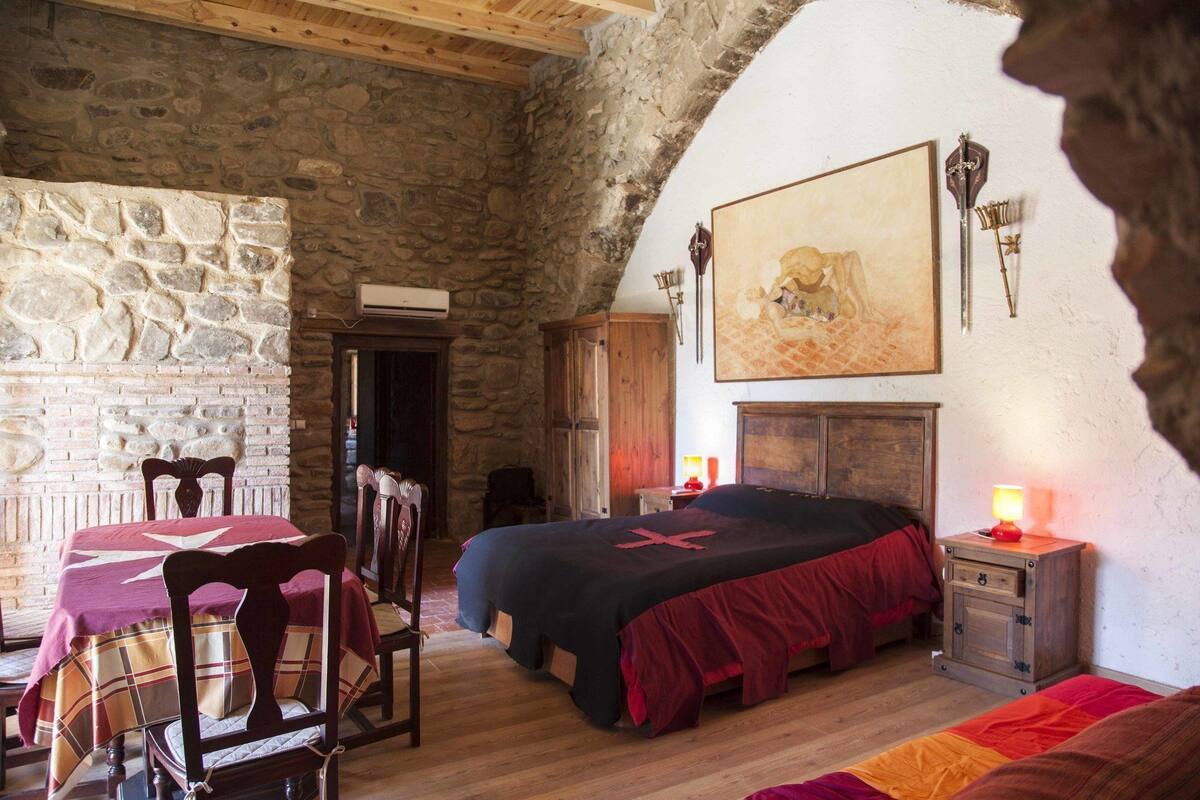 Una habitación rústica con pared de piedra, muebles de madera, una cama con colcha roja y un cuadro en la pared.