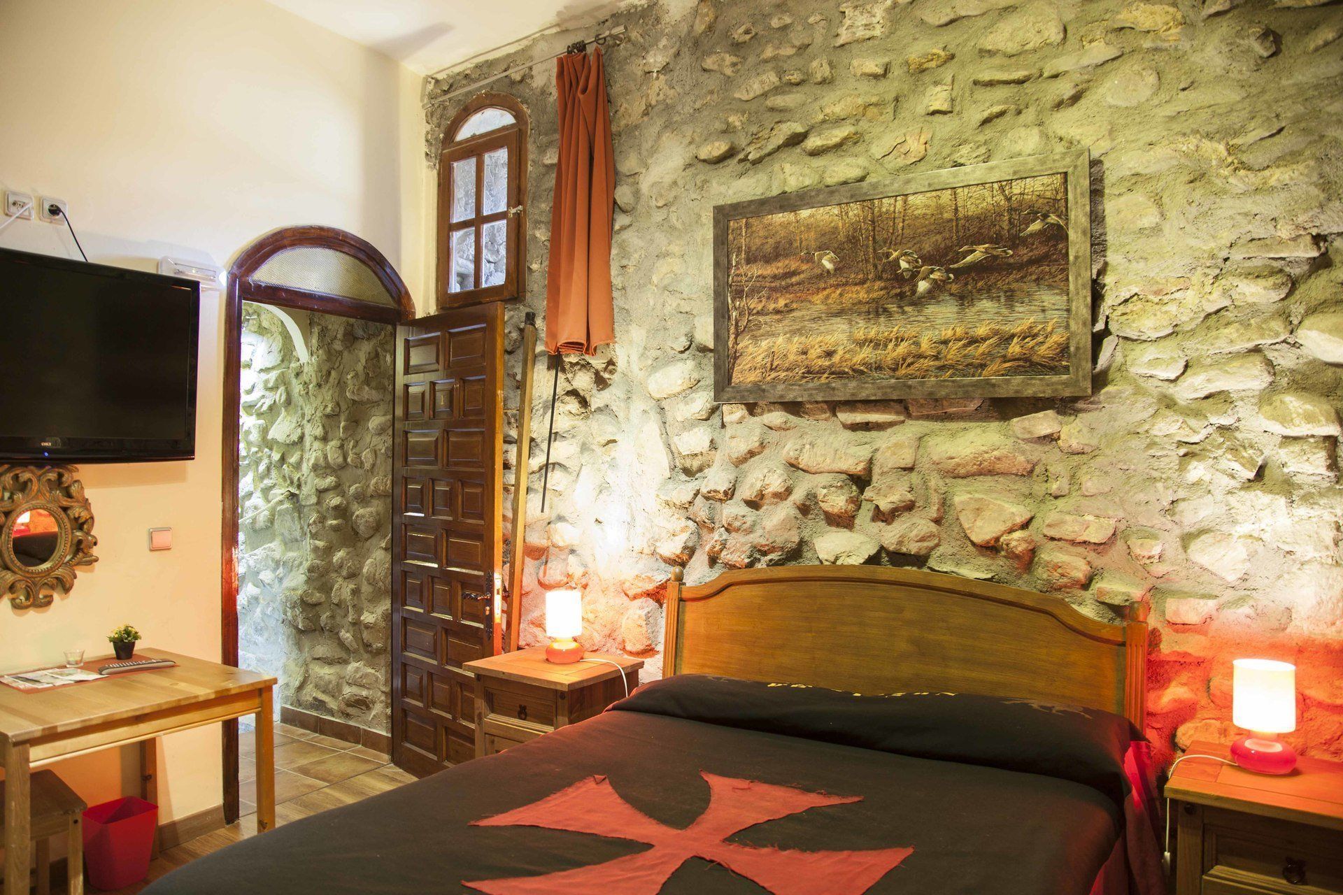 Una camera da letto con parete in pietra, porta in legno, un letto con motivo a croce rossa, un quadro e un televisore appeso al muro.