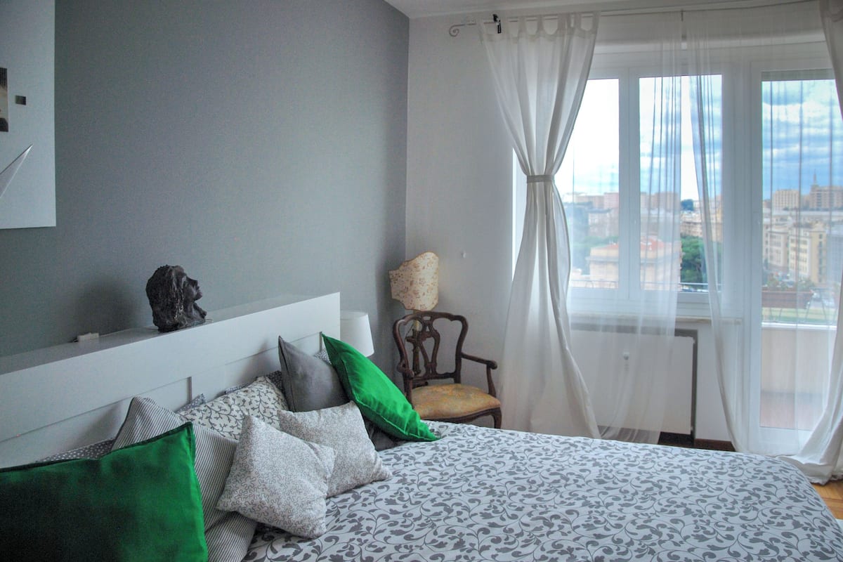 A bedroom with a bed, pillows, a chair, a lamp, and a view of the city through a window with sheer curtains.