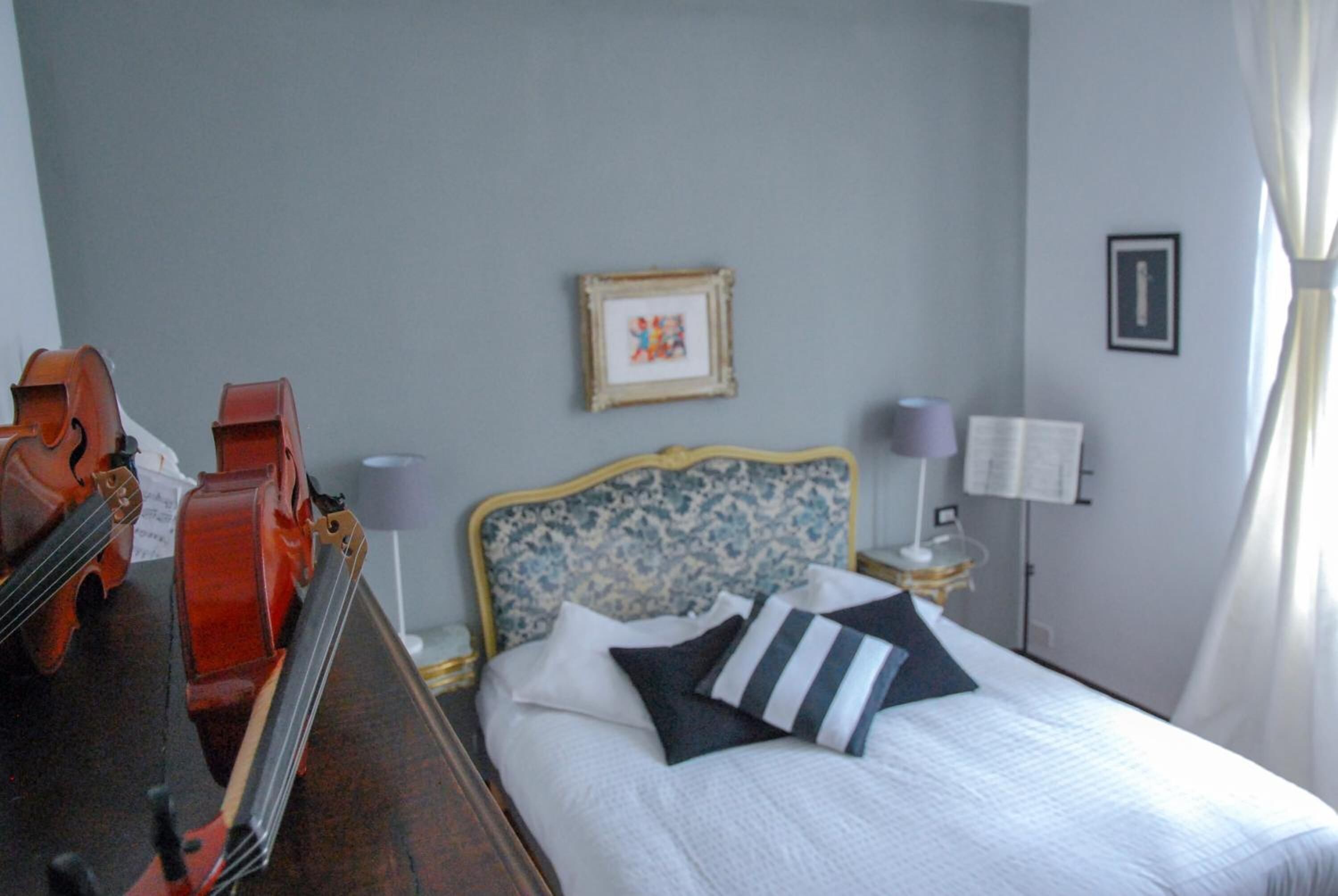 Ein Hotelzimmer mit Bett, Nachttisch, Lampe, einem gerahmten Bild an der Wand und einer Violine auf der linken Seite.