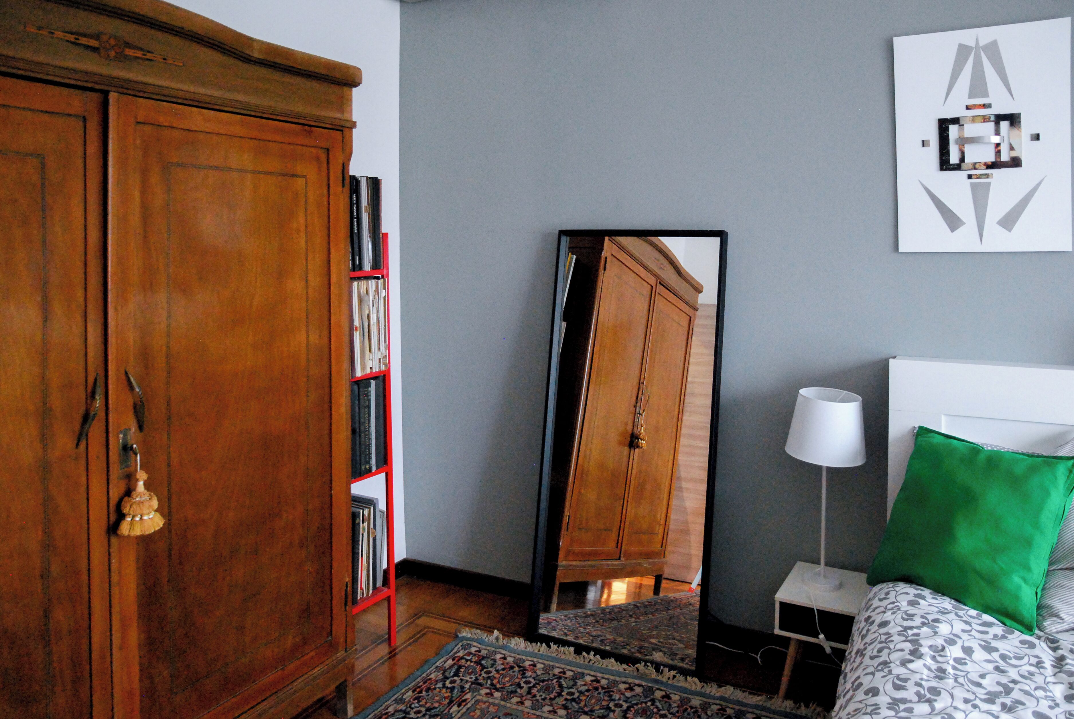 Un dormitorio con un armario de madera, una estantería roja, un espejo grande, una lámpara de noche, un cojín verde y una colcha estampada.