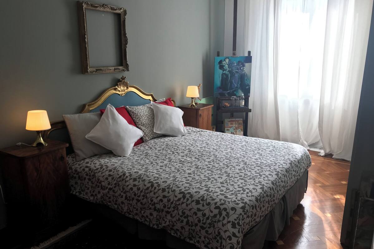 A neatly made bed with patterned bedding, two bedside lamps, a mirror, and a painting on the wall.