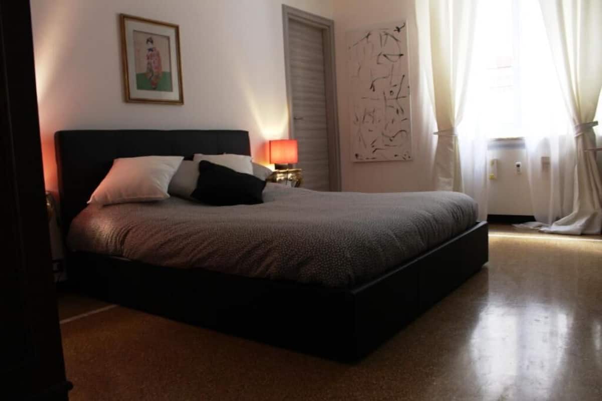 A neatly made bed with a dark headboard, a bedside table with a lamp, a framed picture on the wall, and a large abstract painting.