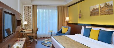 A hotel room with a bed, a desk, a chair, a TV, and a small table with a lamp.