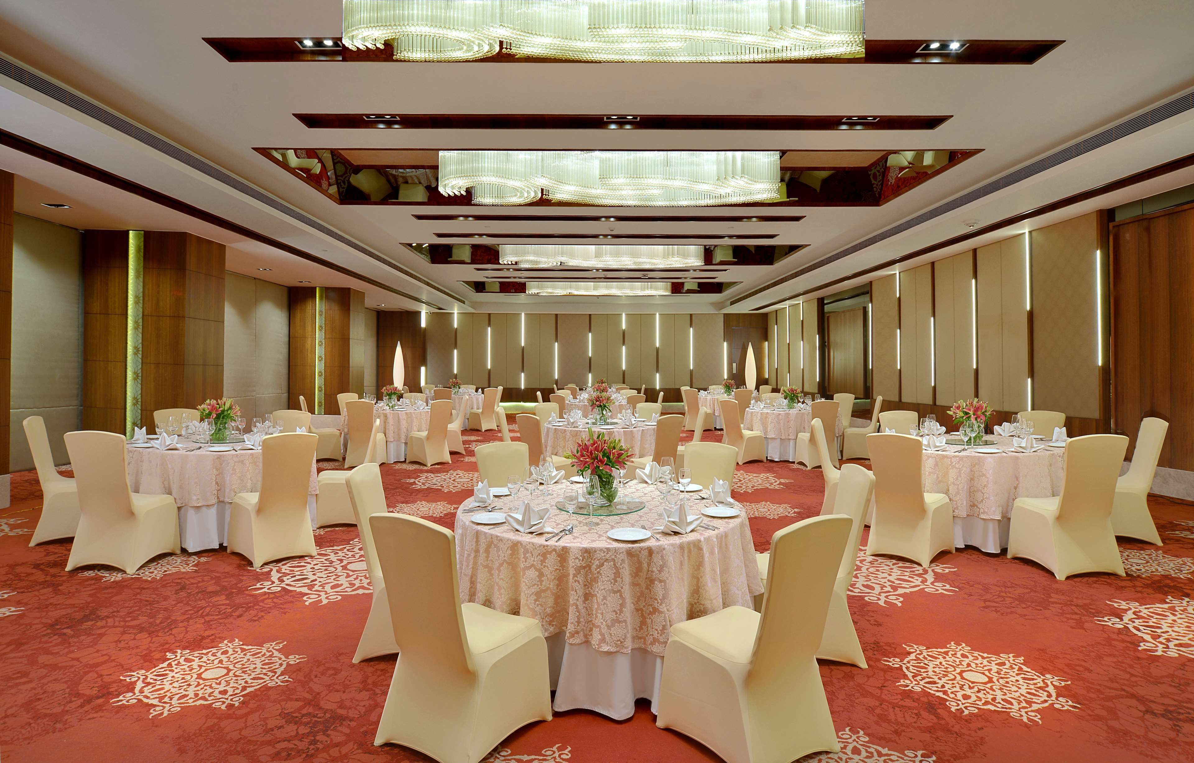 A banquet hall with round tables set for dining, featuring a red carpet and modern chandeliers.