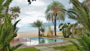 A swimming pool with a wooden deck and stone pathway, surrounded by palm trees and a view of the beach. - Botaniq Hotel Boutique (Mojacar)