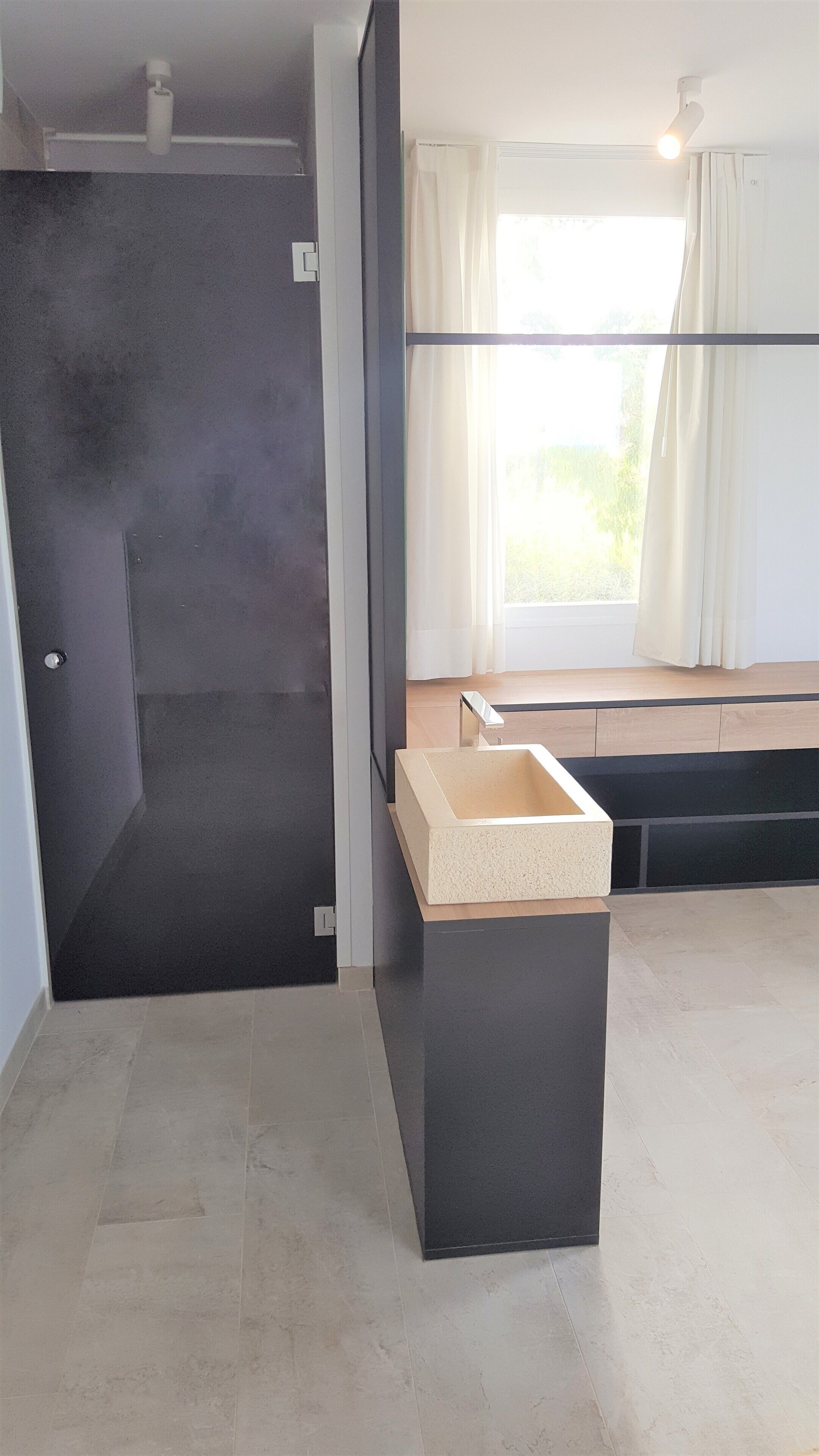 Un baño moderno con piso oscuro, un lavamanos color beige y una ventana con cortinas.