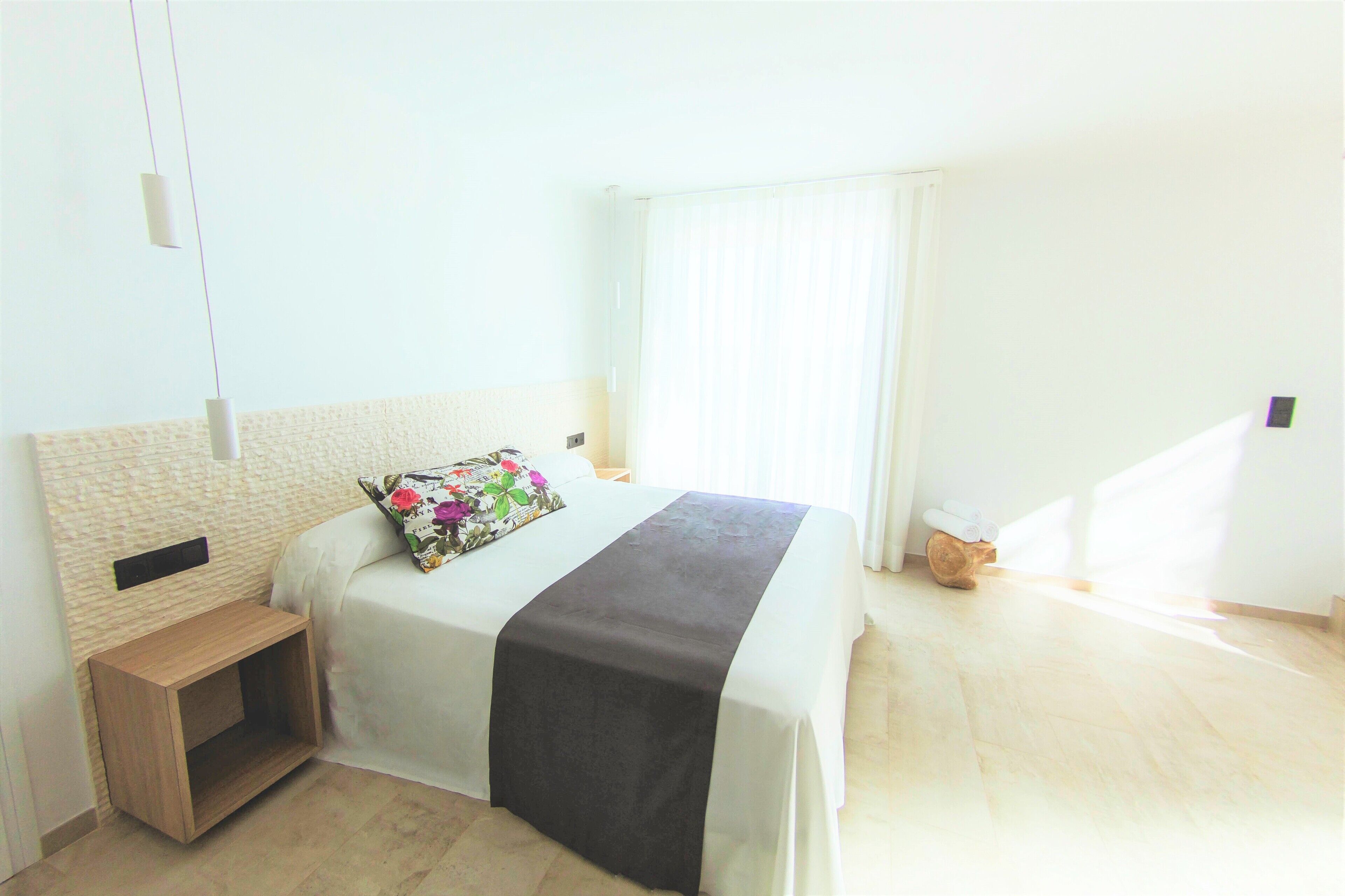 A neatly made bed with a dark bedspread, a wooden nightstand, and a small wooden stool.