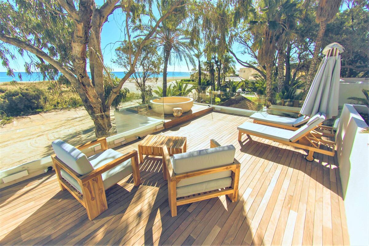A wooden deck with lounge chairs and a sun umbrella, overlooking a beach and palm trees.