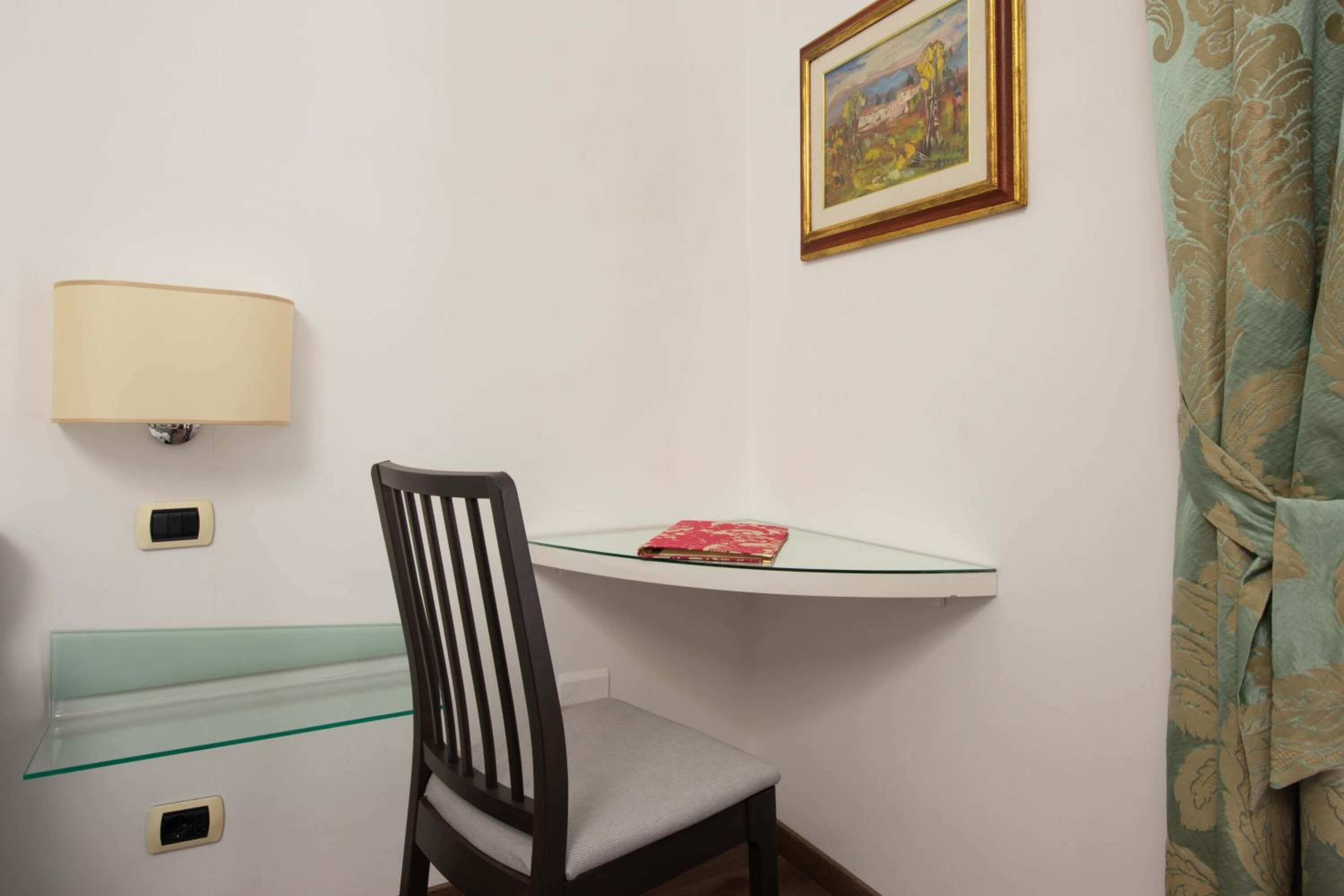 Quarto de canto com mesa de vidro, uma cadeira, luminária fixada na parede e um quadro emoldurado.