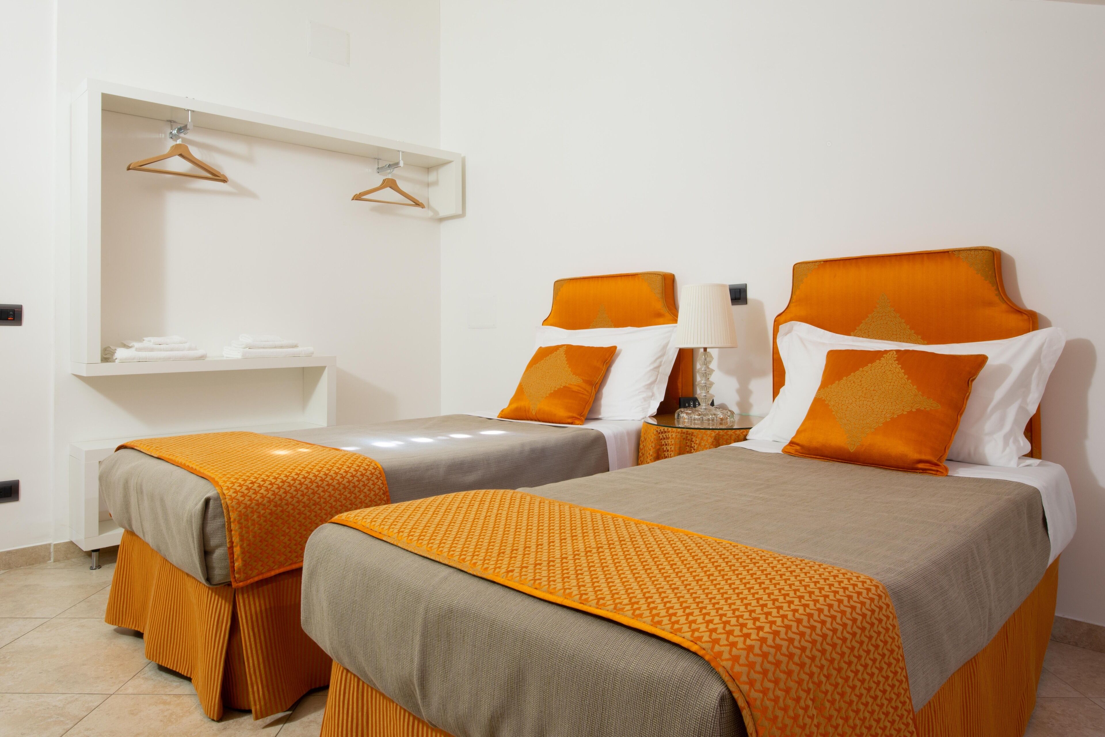 A hotel room with two beds, each with an orange and white bedspread, a bedside table with a lamp, and a white shelf above the beds.