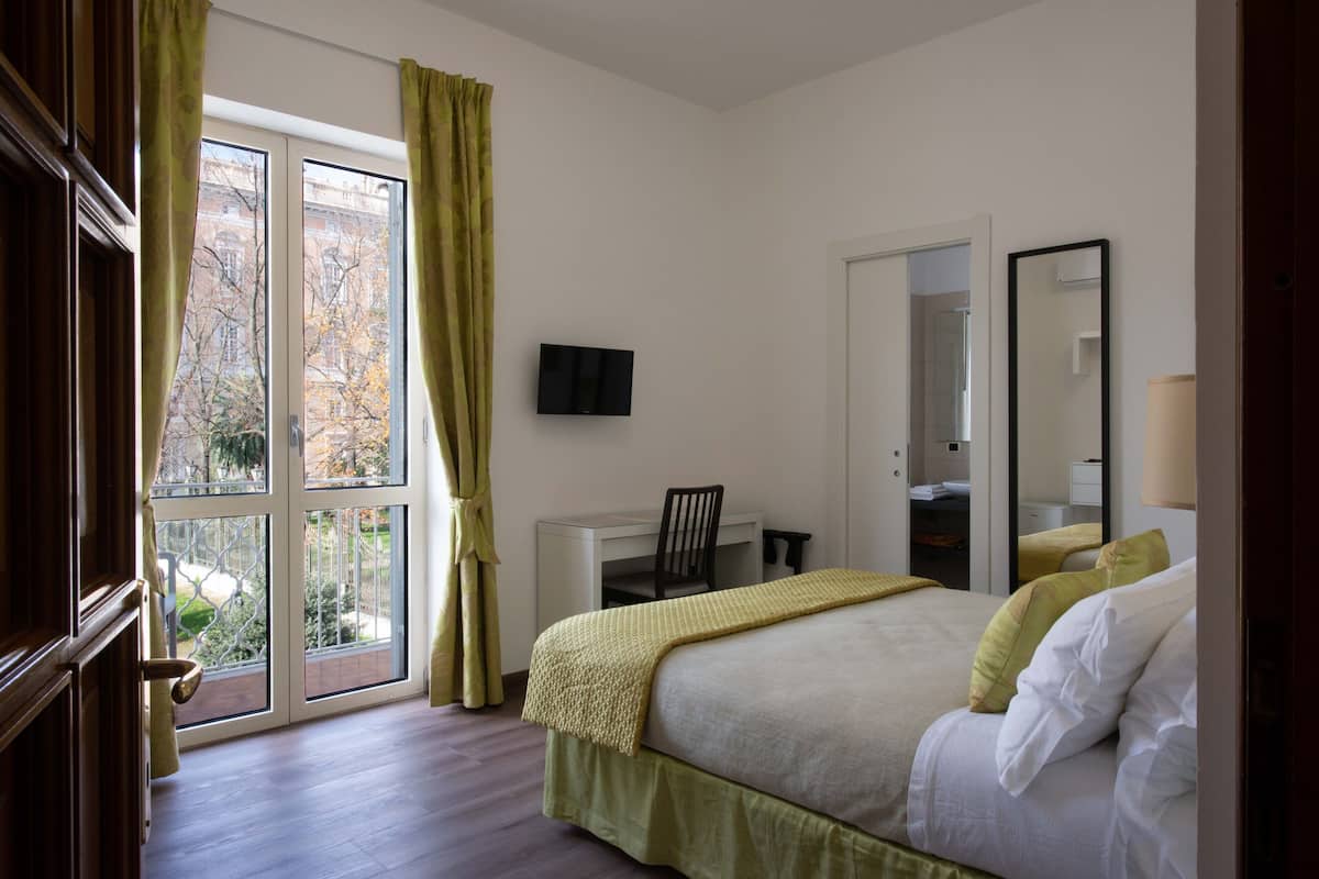 Une chambre à coucher comprenant un lit, un bureau, une chaise, un téléviseur et une grande fenêtre avec des rideaux.