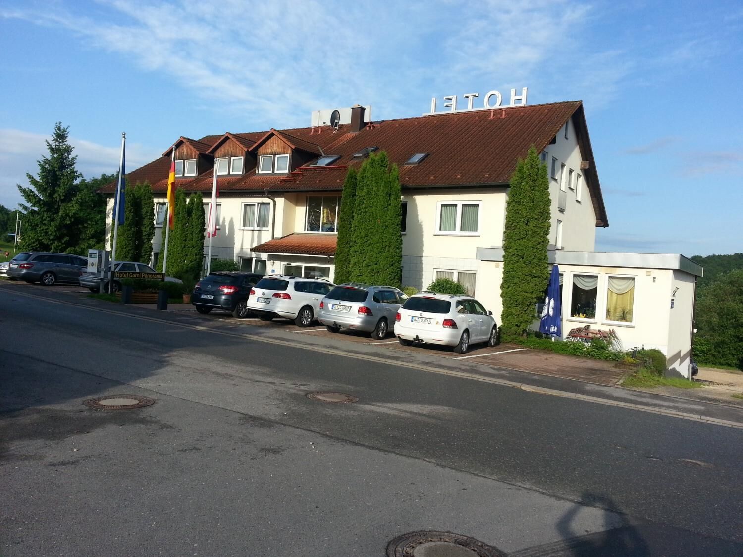 Un hotel dallo stile architettonico europeo tradizionale, con tetto in tegole rosse e numerose finestre. Diverse auto sono parcheggiate davanti e il nome dell'hotel è visibile sull'edificio.