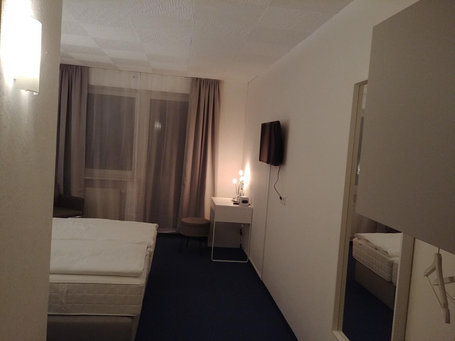 A hotel room with a bed, a desk with a lamp, a TV mounted on the wall, and a window with curtains.