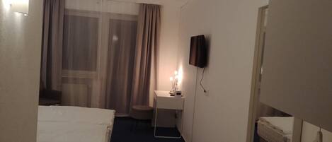 A hotel room with a bed, a desk with a lamp, a TV mounted on the wall, and a window with curtains.