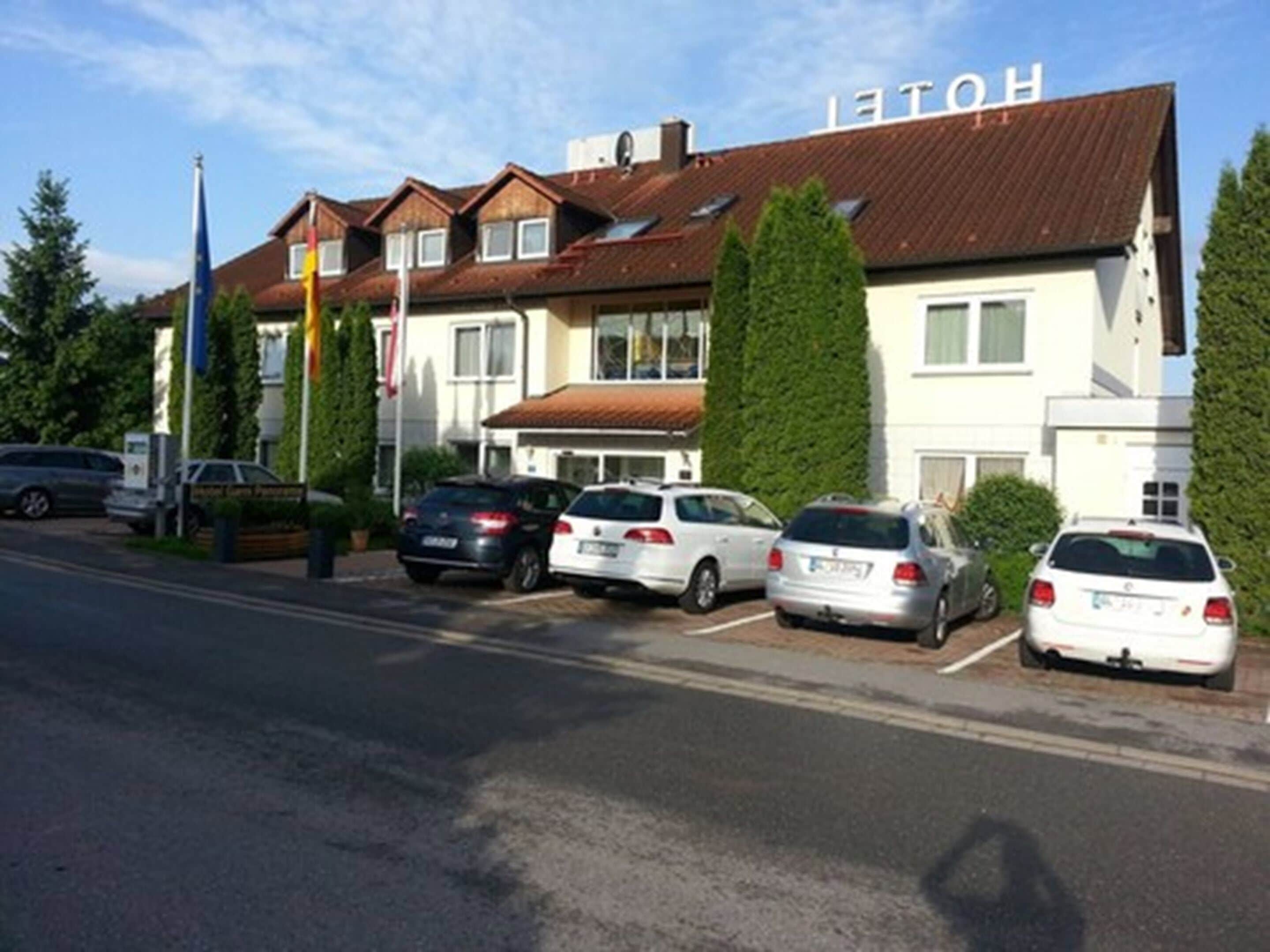 Een hotel in traditioneel Europese stijl, met een bruin tegeldak en witte muren. Er staan meerdere auto’s geparkeerd voor het hotel, en een vlaggenmast met vlaggen is zichtbaar.