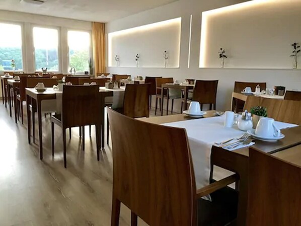 Un restaurant avec des tables et des chaises en bois, des nappes blanches et une vue sur la verdure grâce à de grandes fenêtres.