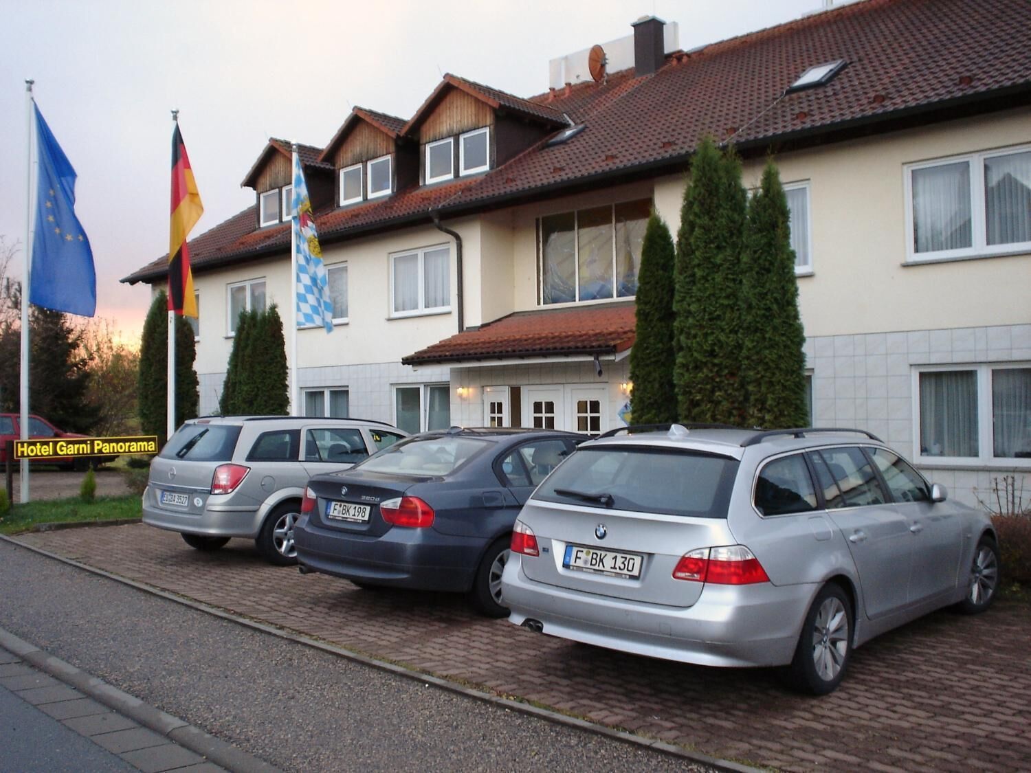 Een rij geparkeerde auto's voor een gebouw met verschillende vlaggen, waaronder de Europese Unie vlag.