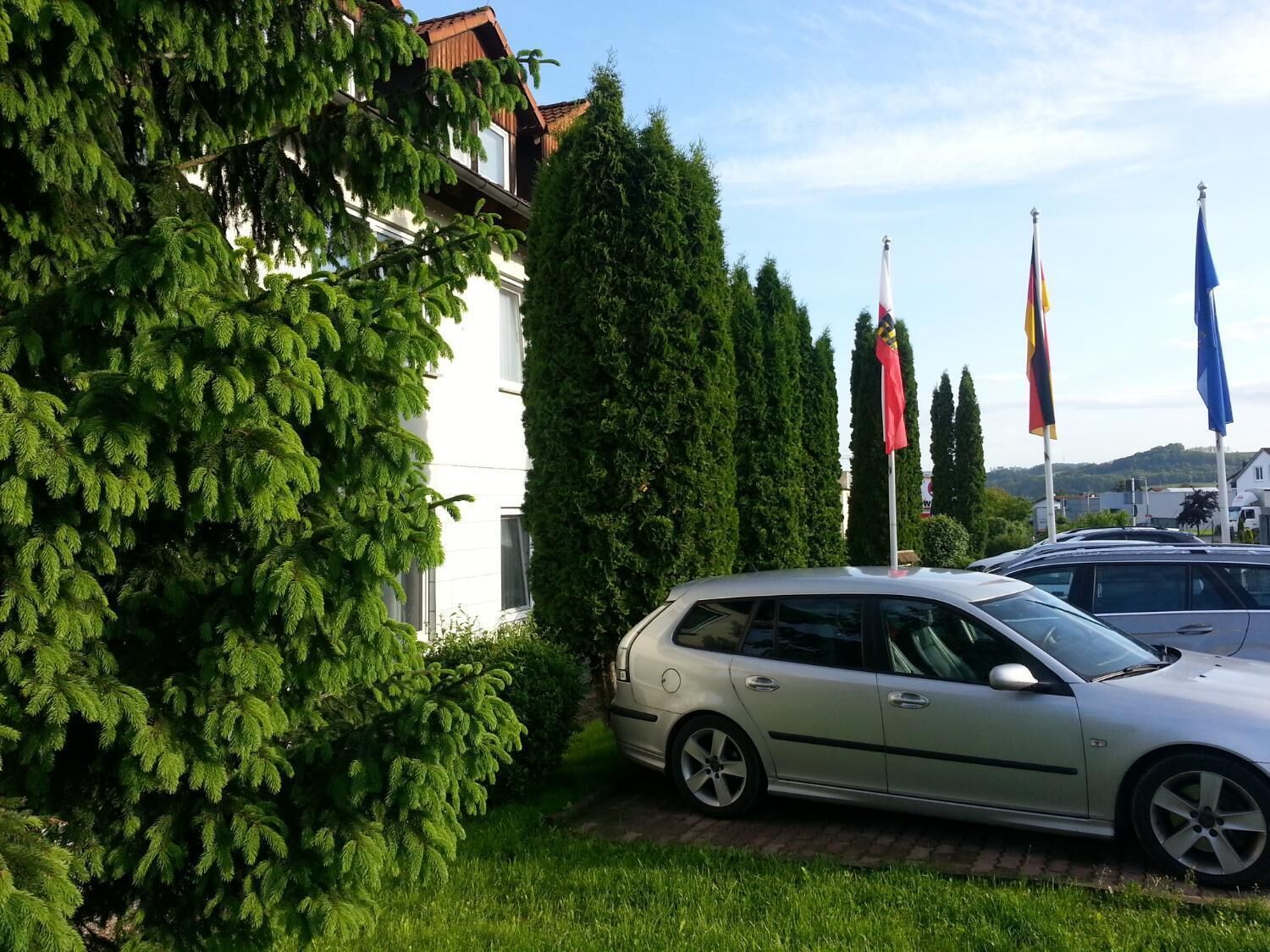 A silver car parked in front of a building with multiple flags, surrounded by greenery.