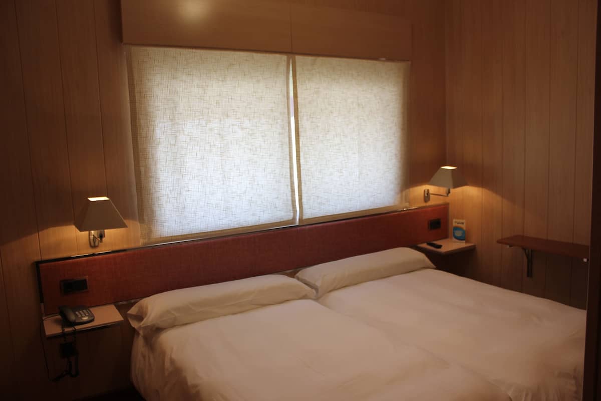 Habitación de hotel con una cama, dos lámparas de noche, una mesita de noche con un teléfono y una ventana con cortinas.