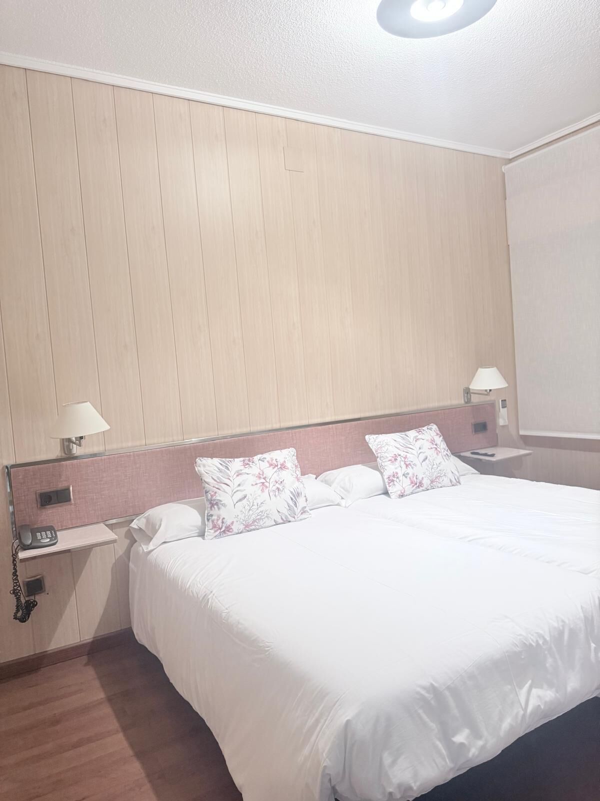 A hotel room with a wooden headboard, a bed with white bedding, two bedside lamps, a phone on the nightstand, and a wall-mounted light fixture.