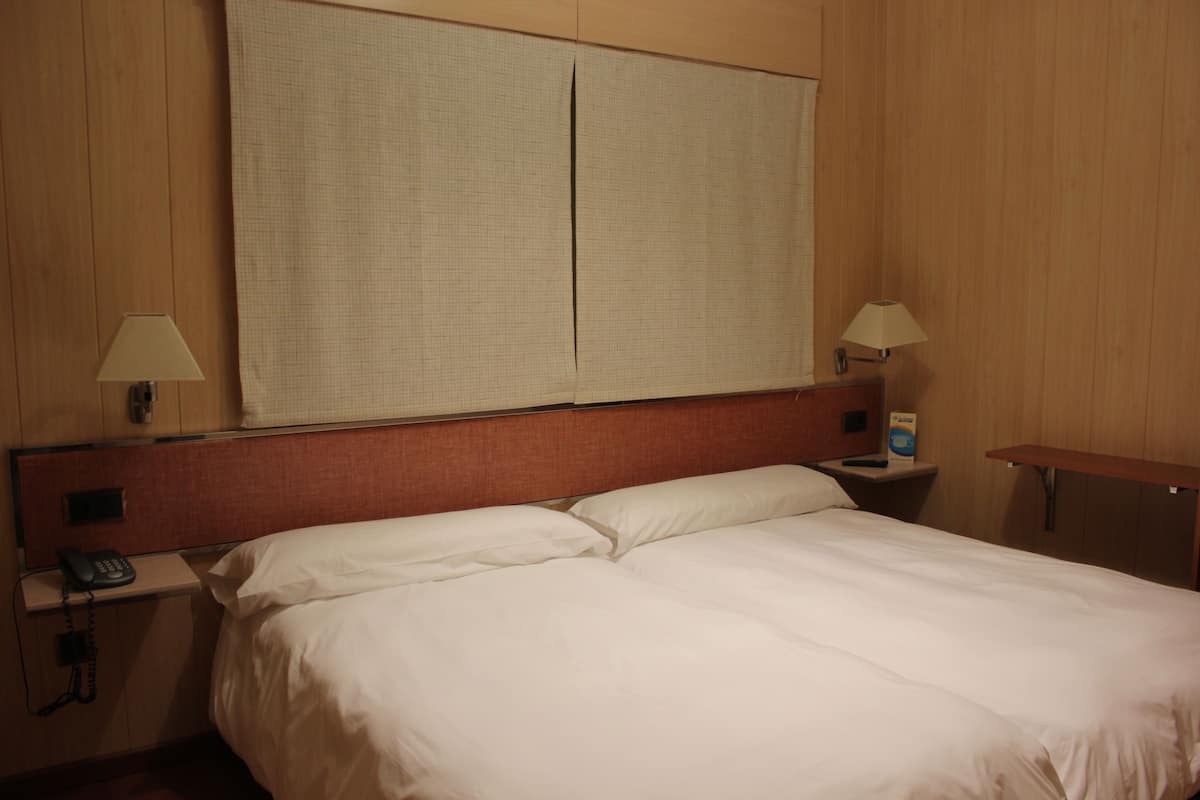Una habitación de hotel con una cama grande, dos mesitas de noche con lámparas y un amplio ventanal con persianas.