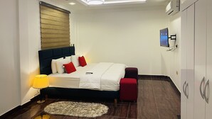 Une chambre à coucher moderne avec un lit, un ventilateur de plafond, un téléviseur fixé au mur et un pouf rouge.