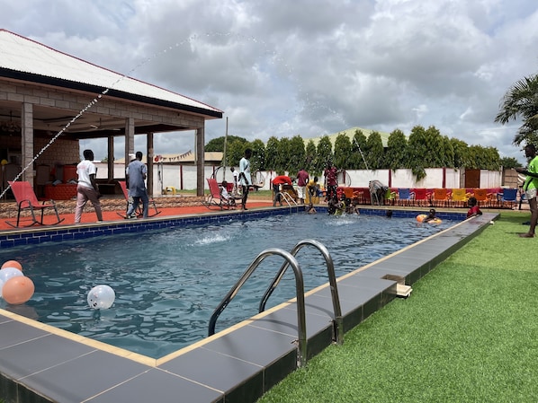 A swimming pool area with a covered seating area, a grassy lawn, and a building with a tiled roof. - Clinton Lodge (Tamale)