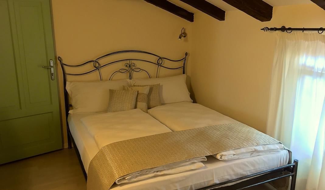 Una cama individual con estructura metálica, una puerta verde y una ventana con cortinas ligeras.