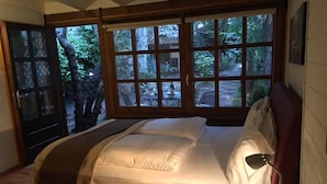 Quarto com uma janela grande, cama com cabeceira vermelha, mesa de cabeceira com luminária e um copo de água.