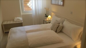 Uma cama de casal com colcha estampada, uma mesinha de cabeceira com abajur e uma pequena planta.