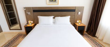 A hotel room with a large bed, two bedside tables, a nightstand, a wall-mounted light, a framed picture, a mirror, and a patterned rug.