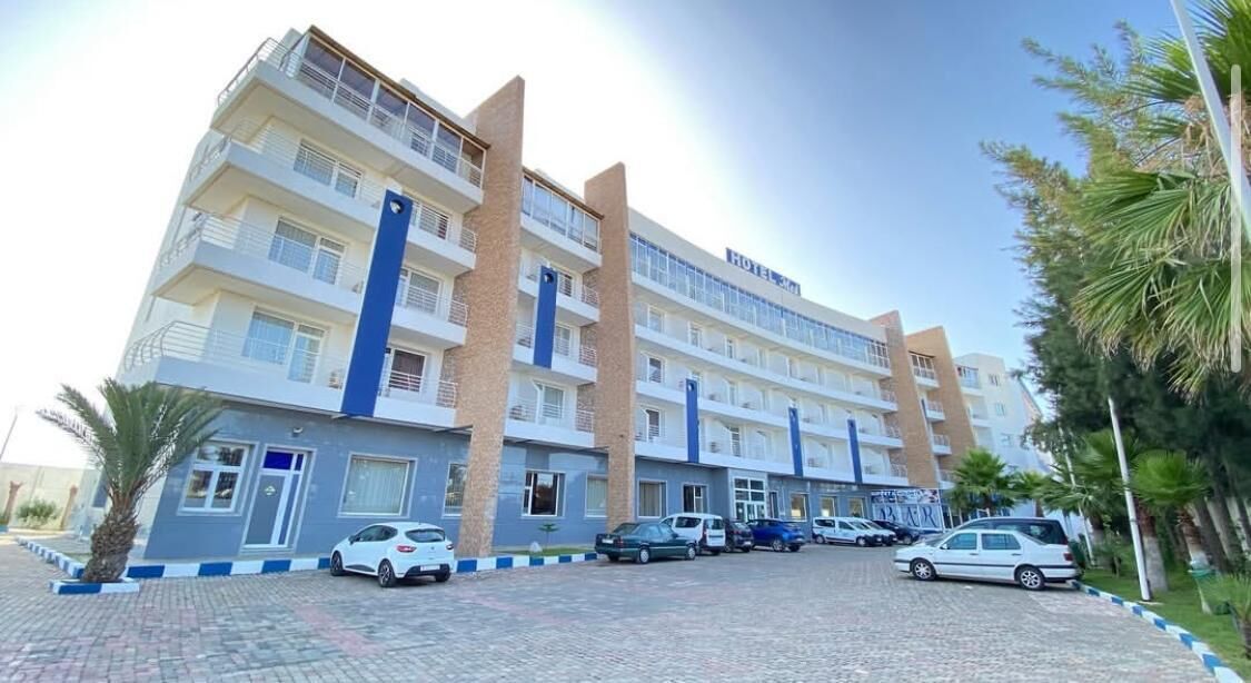 Un hotel de varios pisos con una columna central azul, zona de estacionamiento y palmeras.