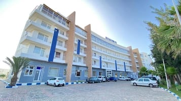 Un hotel de varios pisos con una columna central azul, zona de estacionamiento y palmeras.