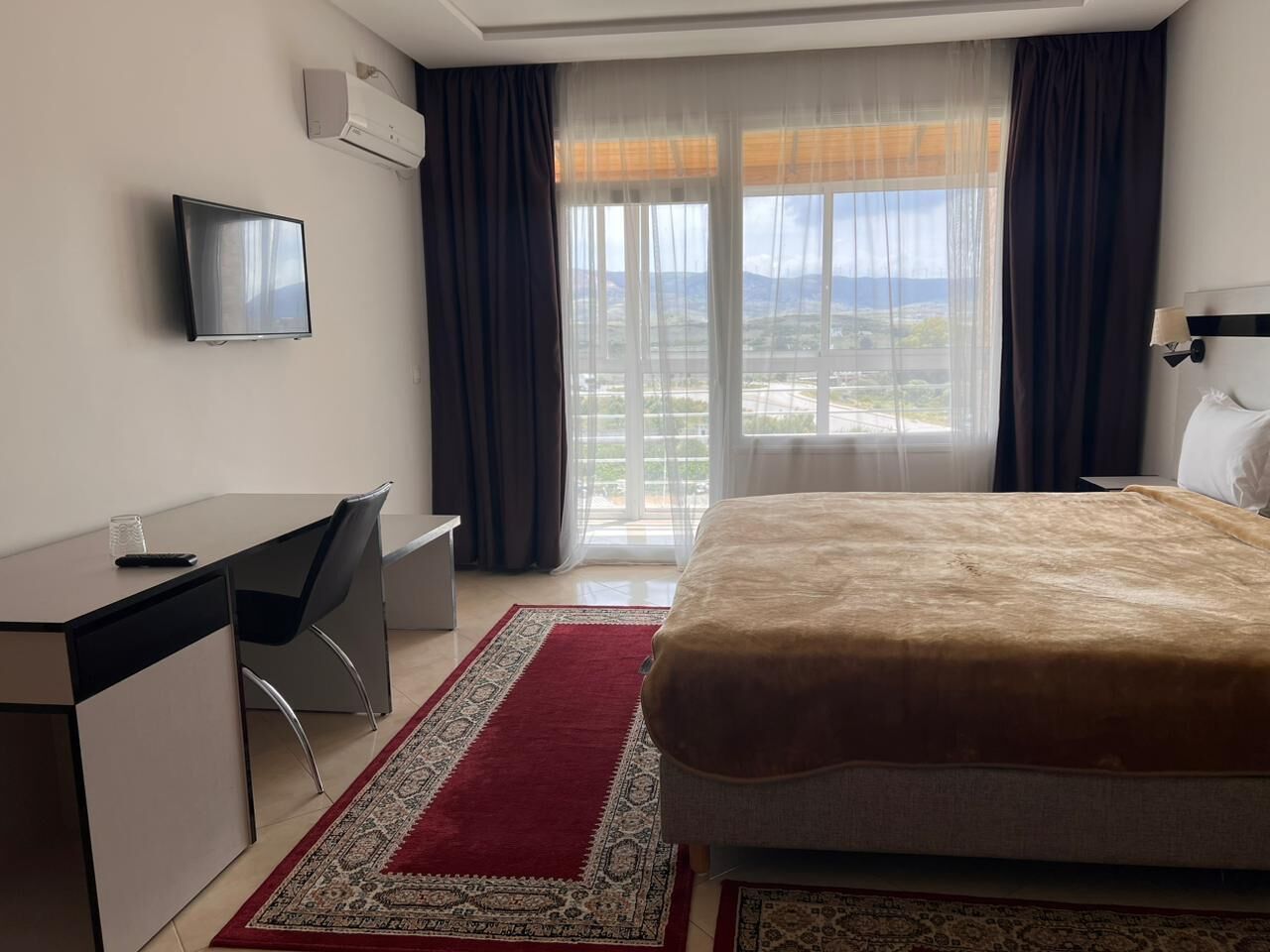 A hotel room with a bed, a desk, a chair, a TV, and a view of the outdoors.