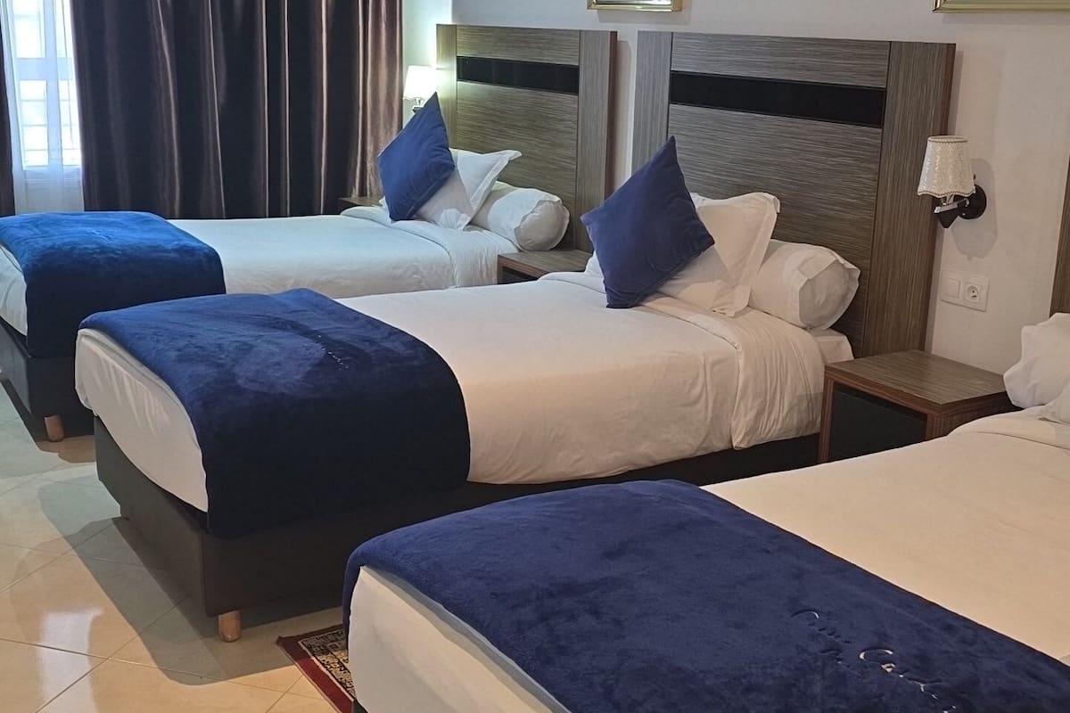 Ein Hotelzimmer mit drei Betten, jedes mit einer blauen Decke und weißen Laken. Zwei wandmontierte Klimaanlagen, ein gerahmtes Bild an der Wand und ein kleiner Tisch mit einer Lampe sind vorhanden.
