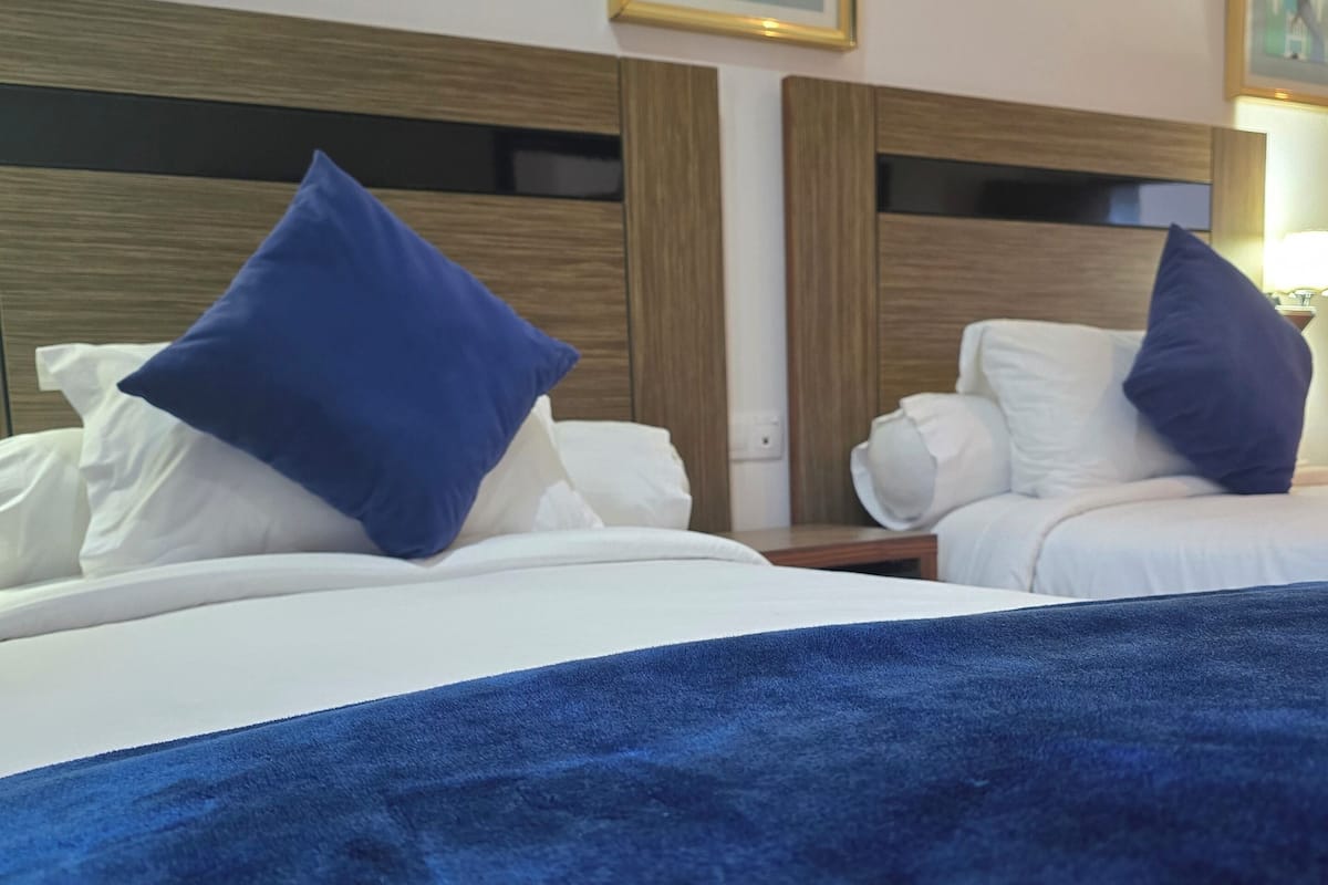 Ein Hotelzimmer mit zwei Betten, jeweils mit weißer Bettwäsche und blauen Kissen. Auf dem Bett im Vordergrund liegt eine große, blaue Decke. Das Zimmer verfügt über hölzerne Kopfteile und ein gerahmtes Bild an der Wand.