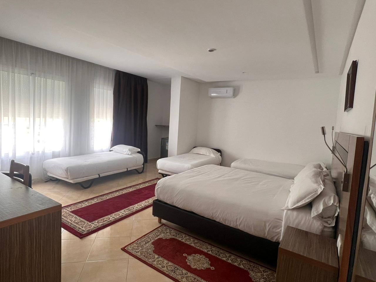 Une chambre d’hôtel avec deux lits simples, un bureau, une chaise, un tapis aux motifs rouges et beiges, et un climatiseur fixé au mur.