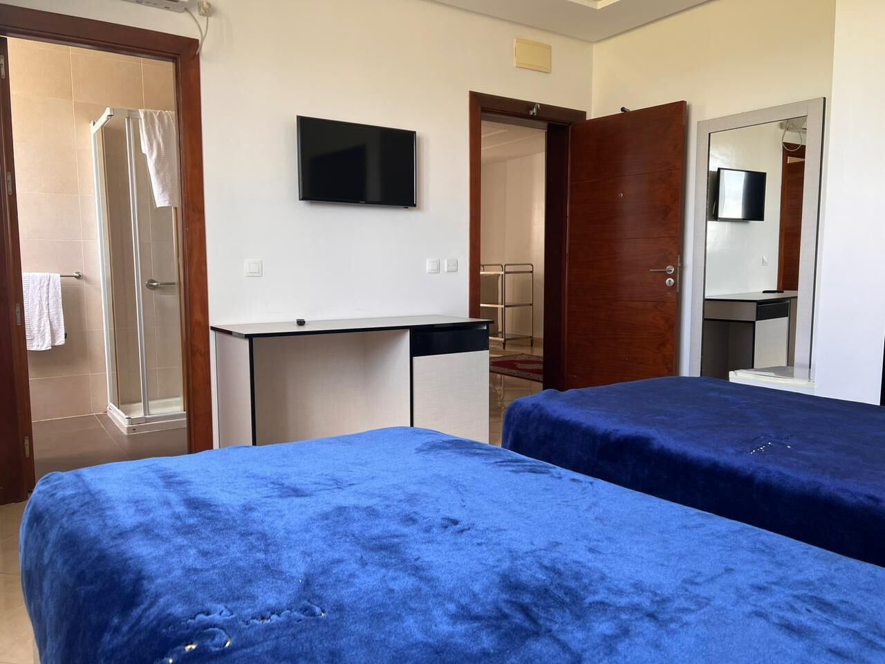 A room with two beds covered in blue bedding, a small desk, a TV on the wall, and a shower area.