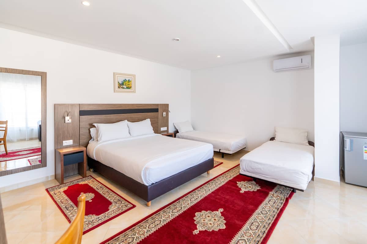 Ein Hotelzimmer mit einem großen Bett, zwei Einzelbetten, einem Spiegel, einem Bild an der Wand und einem Klimagerät, sowie einem roten Teppich.
